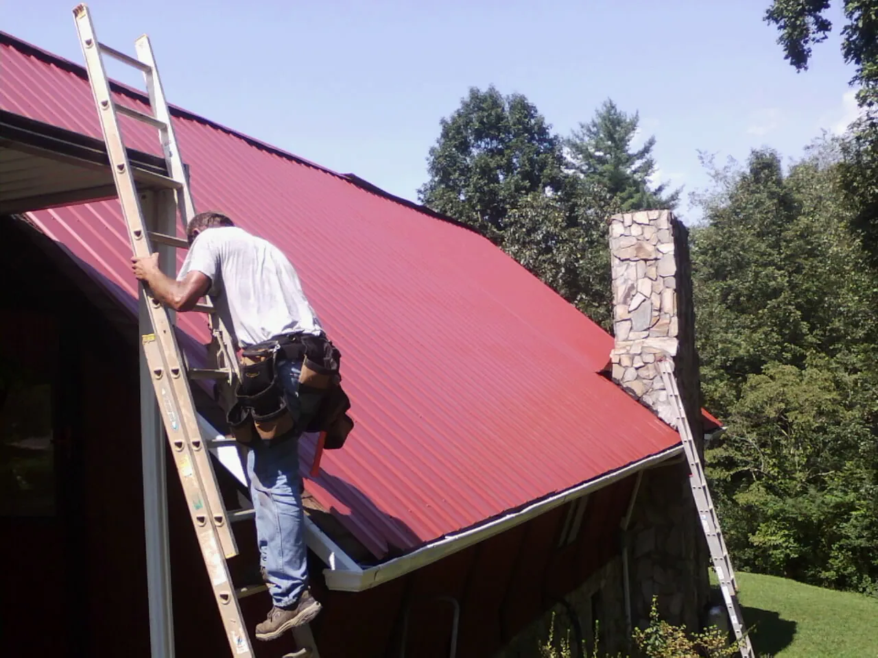 Roofer on a ladder beside a red metal roof and stone chimney — Soffit & Fascia Repair by Roofing Brothers in Grampian